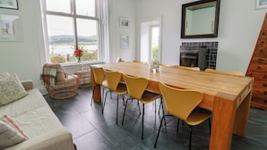 Ein Essbereich mit Holztisch und gelben Stühlen, einem Korbstuhl, einer Couch mit gemusterten Kissen und Blick ins Freie durch ein großes Fenster.
