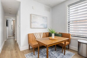 Un coin repas avec une table en bois et deux chaises, une plante en pot et une fenêtre avec des stores.