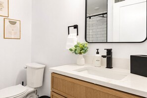 Une salle de bain moderne avec un lavabo blanc, un robinet noir et un miroir au-dessus. Il y a des toilettes à gauche et une poubelle en dessous.