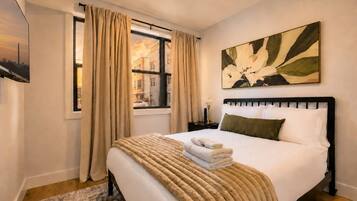 A hotel room with a bed, a window with curtains, a painting on the wall, and a nightstand with a lamp.
