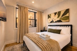 Quarto de hotel com cama, janela com cortinas, pintura na parede e mesa de cabeceira com abajur.