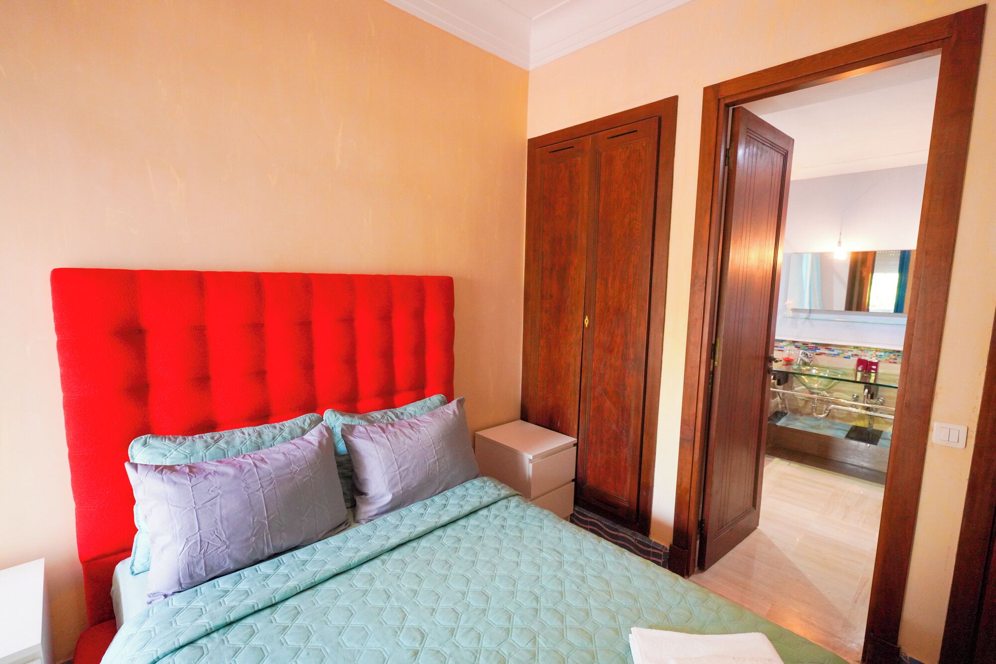 Une chambre à coucher avec une tête de lit rembourrée de tissu rouge, un lit recouvert d’une courtepointe bleu clair et une armoire en bois.