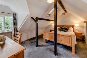 Ein Schlafzimmer mit schrägem Dach, einem hölzernen Bett, einem Nachttisch, einer Lampe und einem Fenster mit Blick auf Grünflächen.