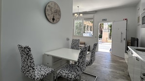 Uma área de refeições com uma mesa branca e cadeiras estampadas, um relógio na parede e vista para o exterior.