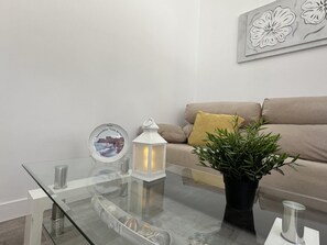 Una sala de estar moderna con una mesa de centro de vidrio, un farol blanco, un cuadro enmarcado, una planta en maceta y un sofá color beige.