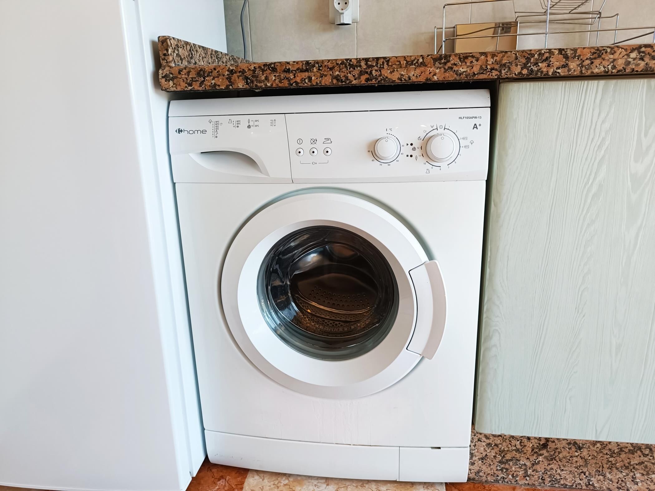 Un lave-linge à chargement frontal doté d’un panneau de commande, placé devant un plan de travail de cuisine avec un pare-feu en granit.