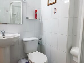 Ein Badezimmer mit einem weißen Waschbecken, einer Toilette und einer Dusche. Über dem Waschbecken hängt ein Spiegel und darunter befindet sich ein Ablagefach mit Toilettenartikeln.