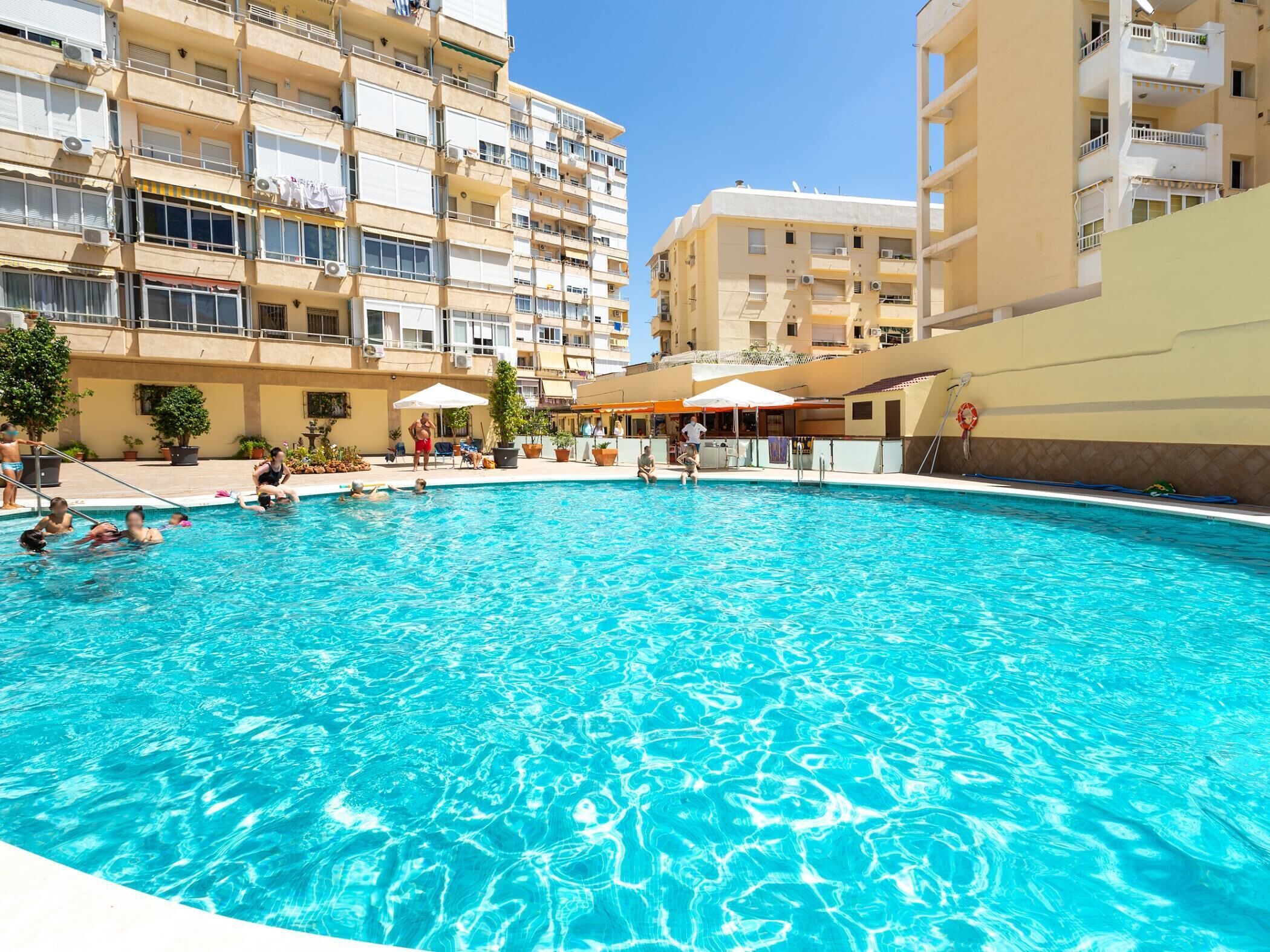 Una piscina rodeada de edificios de varios pisos con balcones, mobiliario exterior y plantas en macetas.