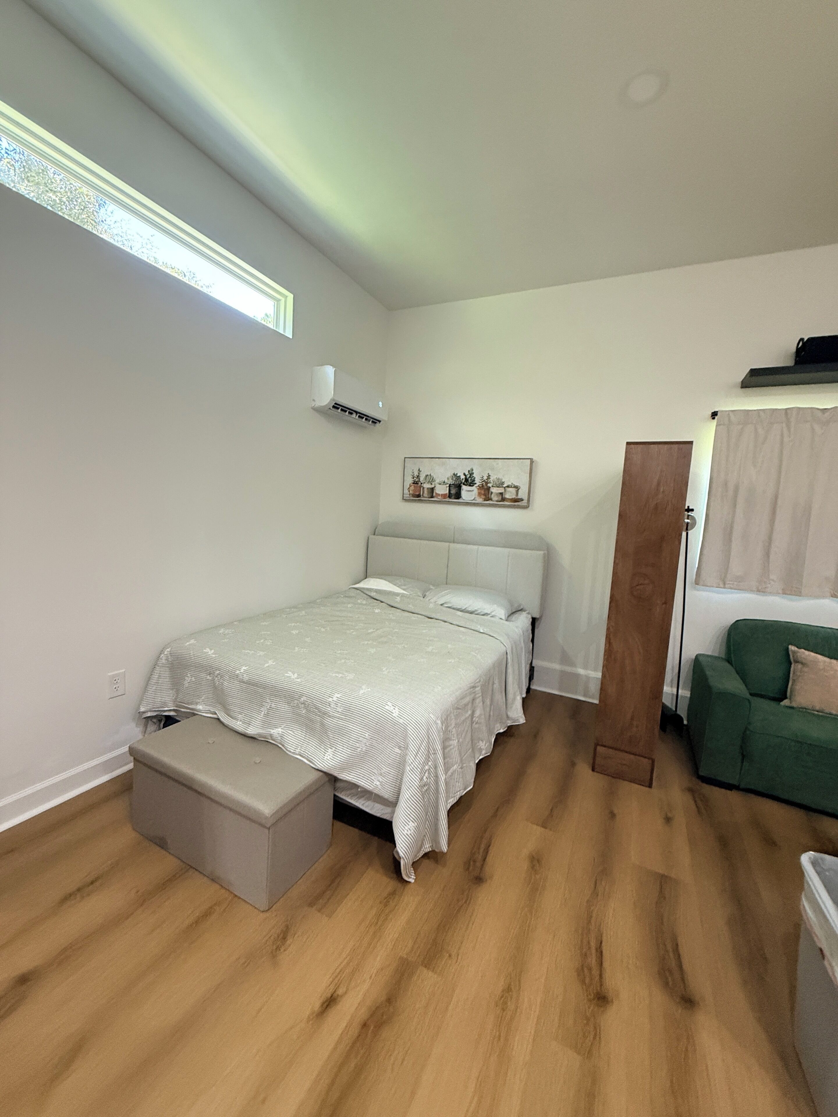A bedroom with a bed, a bench, a green sofa, a wooden wardrobe, and a wall-mounted air conditioner.
