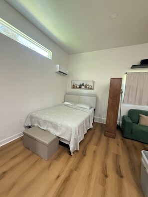 Ein Schlafzimmer mit einem Bett, einer Bank, einem grünen Sofa, einem Holzschrank und einer wandmontierten Klimaanlage.
