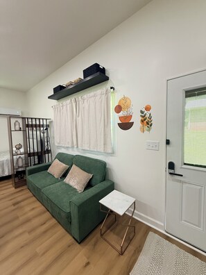 A living room with a green sofa, a small white table, a door with a window, and a wall-mounted shelf. - The R&R Studio  Cozy Private Guesthouse (Fayetteville)