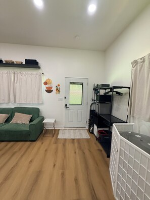 Ein Zimmer mit einem grünen Sofa, einer weißen Tür, einer weißen Wand, einem Regal mit Körben und einem weißen Heizkörper.