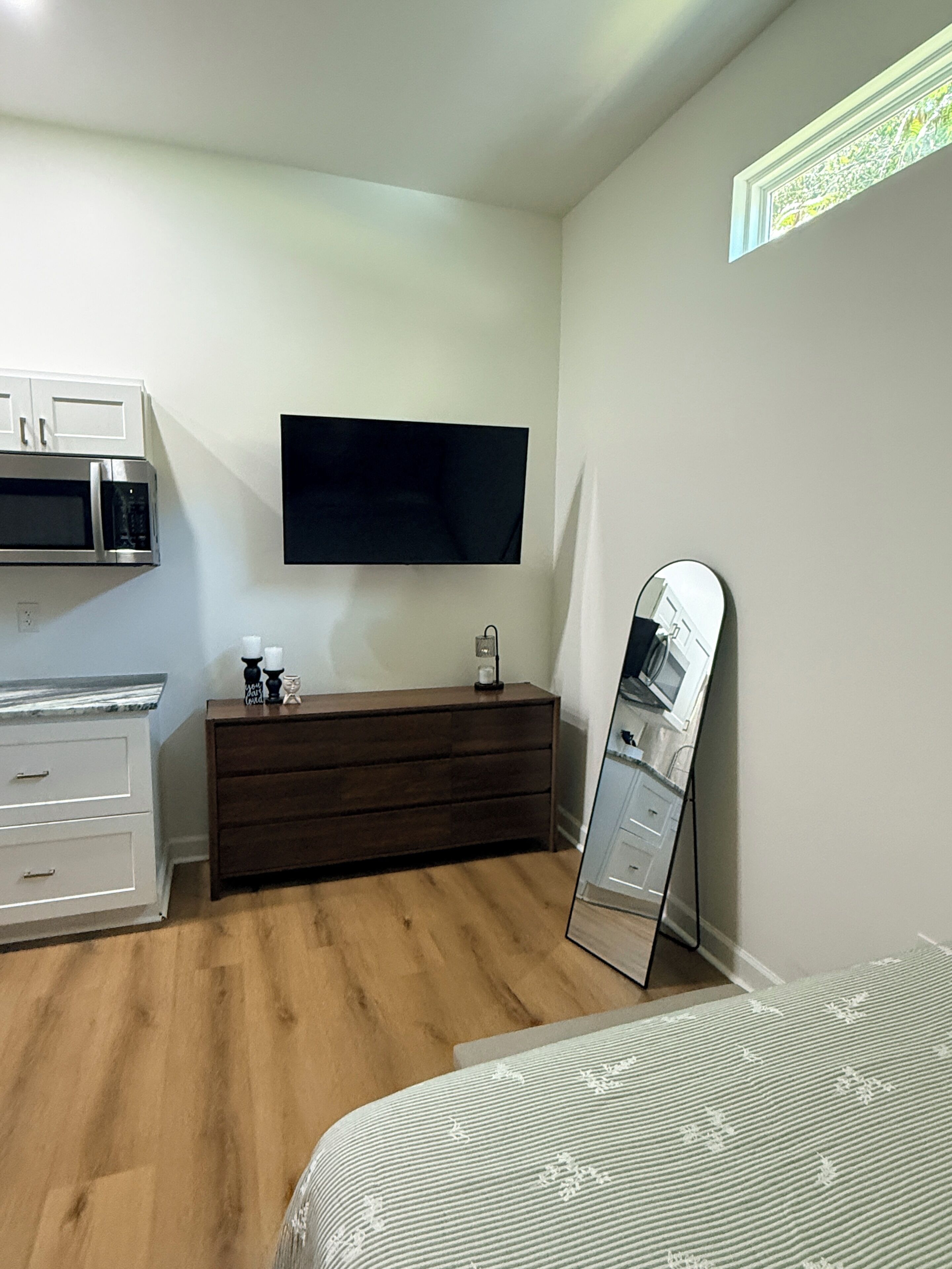 Un dormitorio con piso de madera, cama, cómoda, espejo, televisor de pantalla plana y microondas.