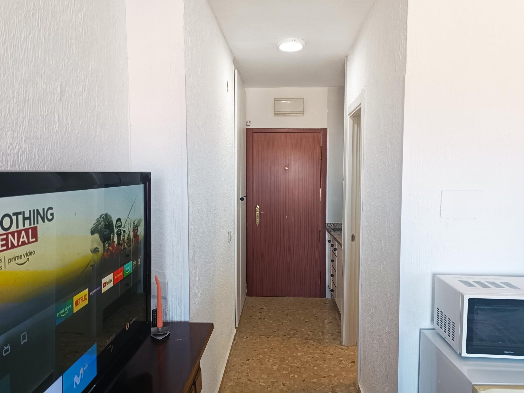 Un pasillo con un televisor a la izquierda mostrando un cartel de película, un microondas a la derecha y una puerta de madera al final.