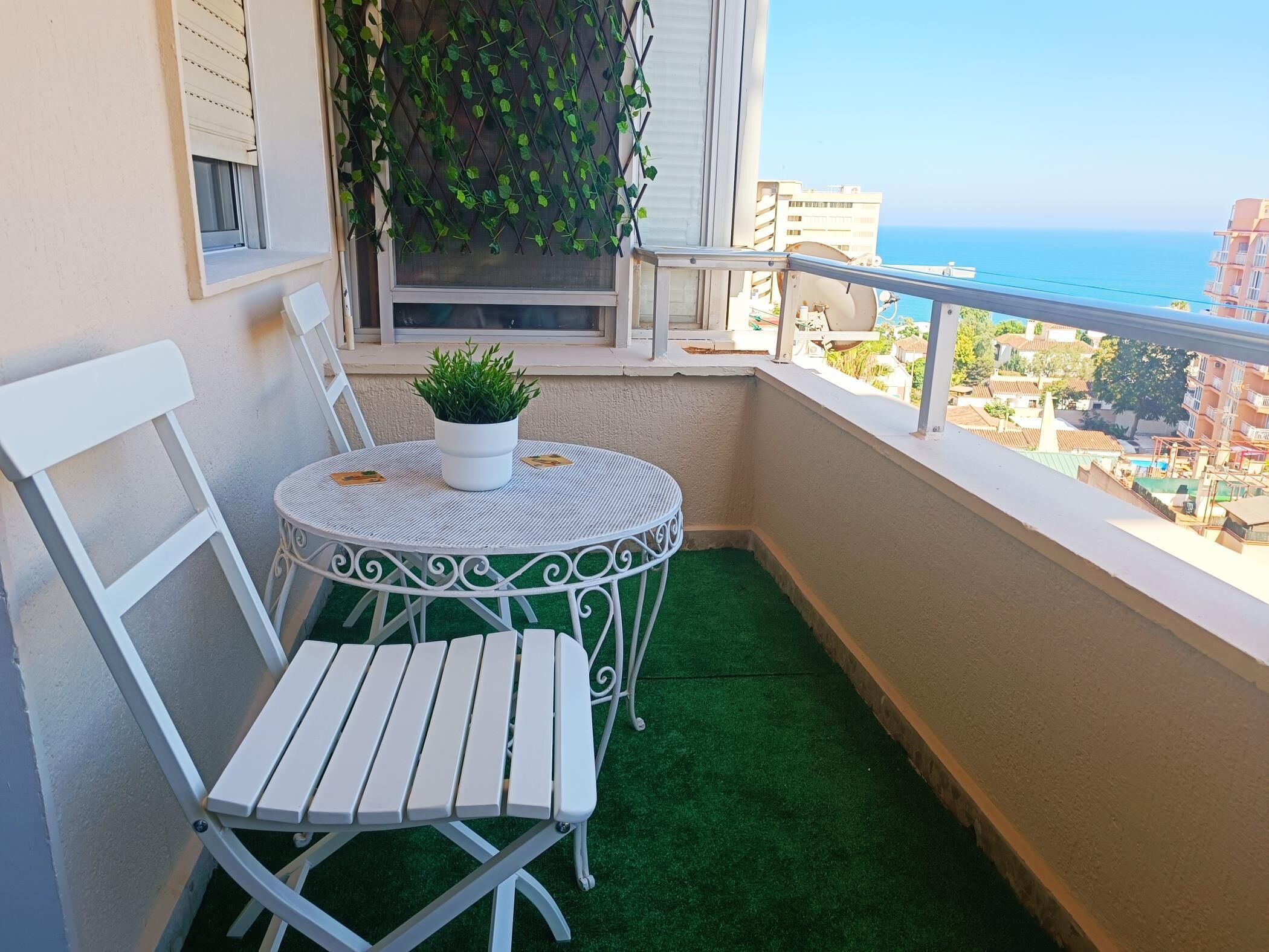 Un balcon avec une petite table et deux chaises, un pot de fleurs et une vue sur la mer et les bâtiments.