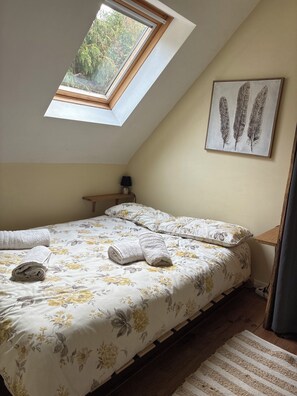 A bedroom with a skylight, a bed with floral bedding, a nightstand, and a wall-mounted feather artwork. - Fishermen's Cottage (Pluméliau-Bieuzy)
