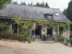 Ein Steinhaus mit einem geneigten Dach, einer Haustür und einer kleinen Veranda mit Sitzgelegenheiten.