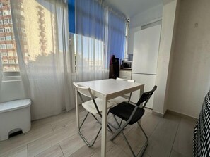Een knus woongedeelte met kitchenette, een eethoek met tafel en stoelen, en uitzicht naar buiten door een raam met transparante gordijnen.