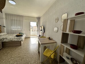 Une chambre à coucher avec un lit, un bureau et une chaise. Il y a une fenêtre avec des rideaux, un téléviseur fixé au mur et des étagères ornées d’objets décoratifs.