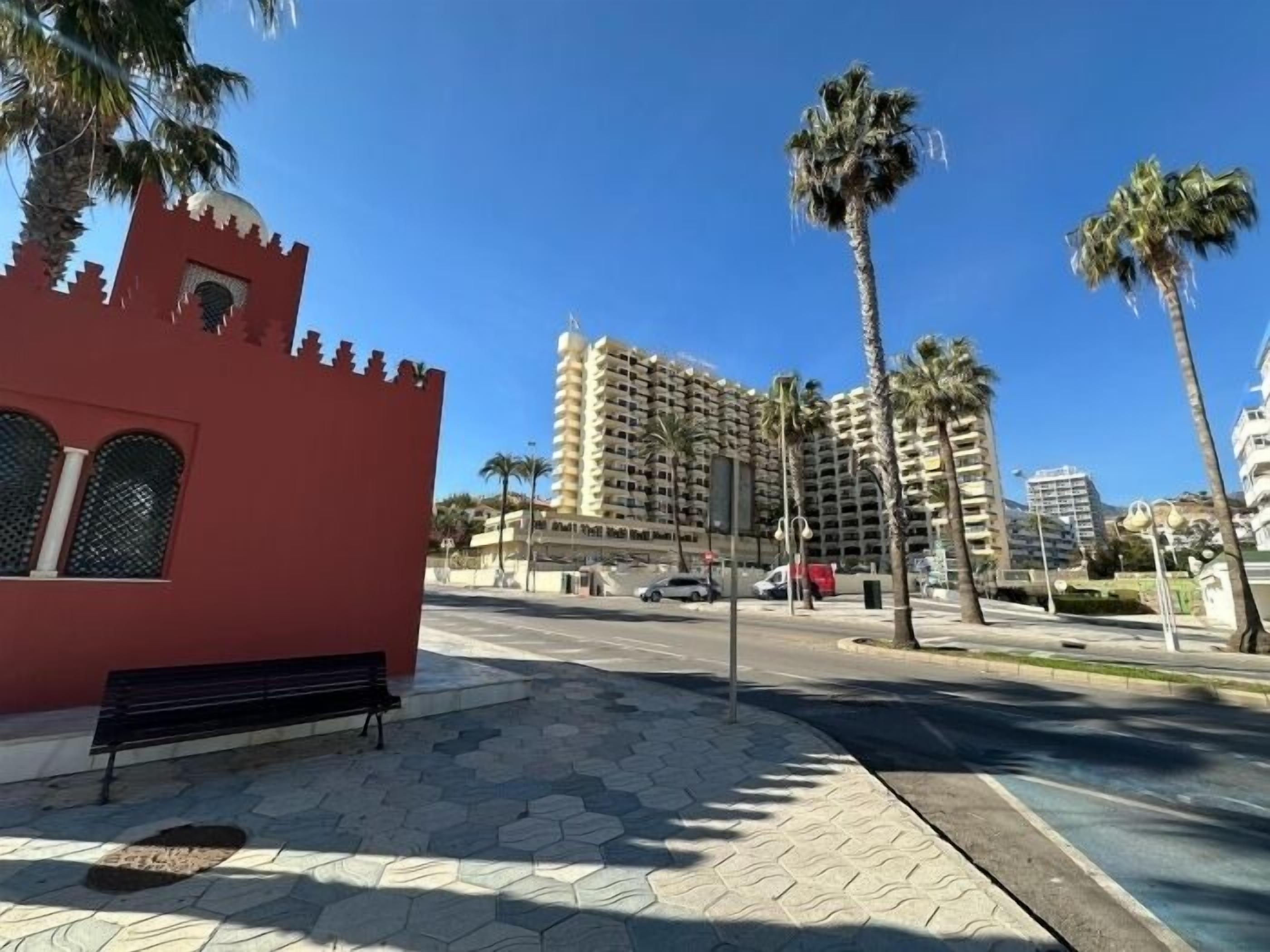 Uma passarela pavimentada com um prédio vermelho, palmeiras e um hotel de vários andares ao fundo.
