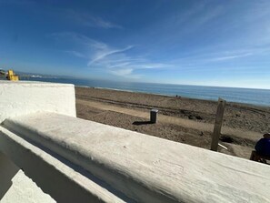 Blick auf einen Strand vom Balkon aus, mit Geländer, blauem Himmel und einer fernen Küstenlinie.