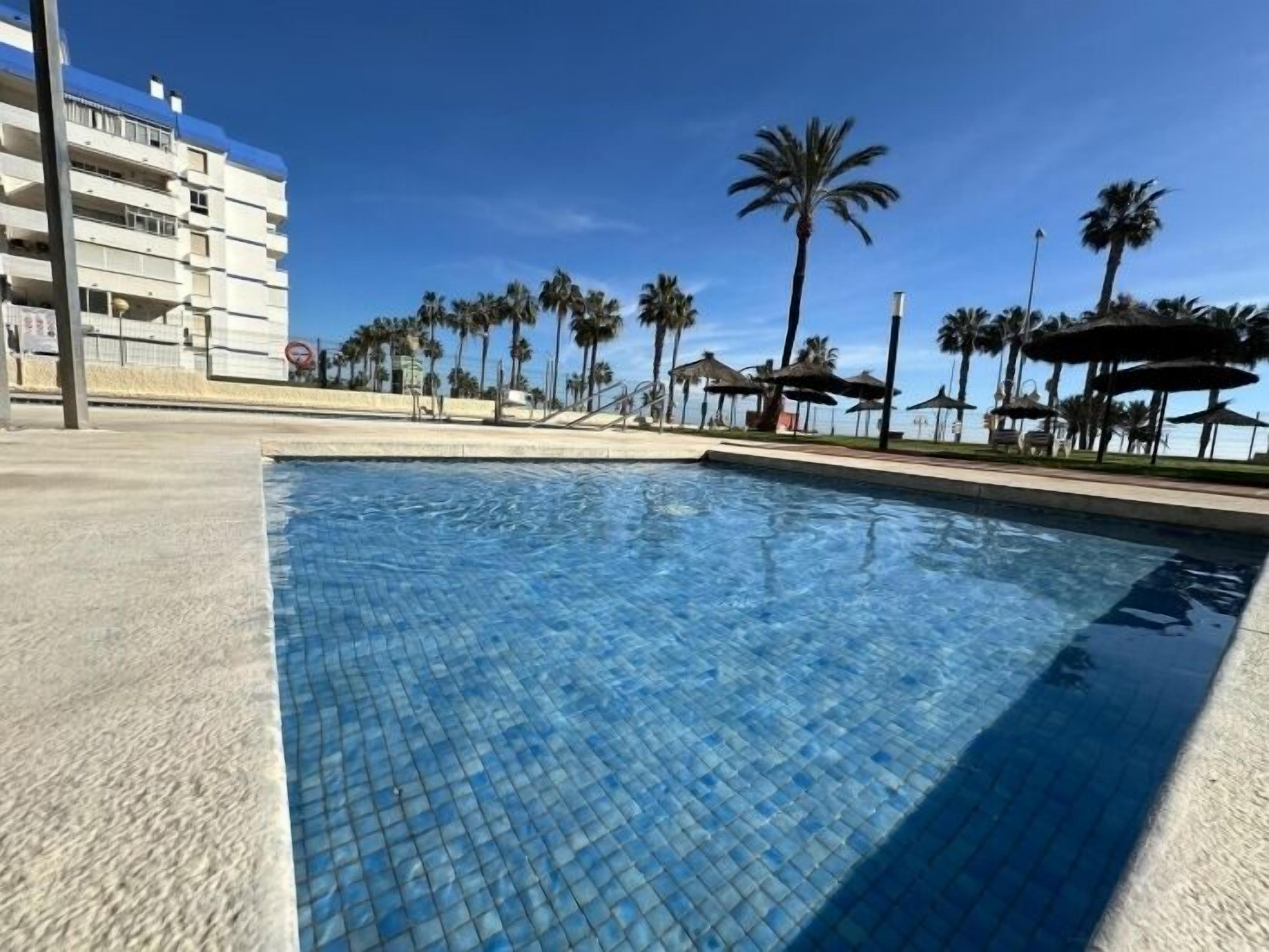 A swimming pool with tiled flooring, surrounded by palm trees and a building in the background.