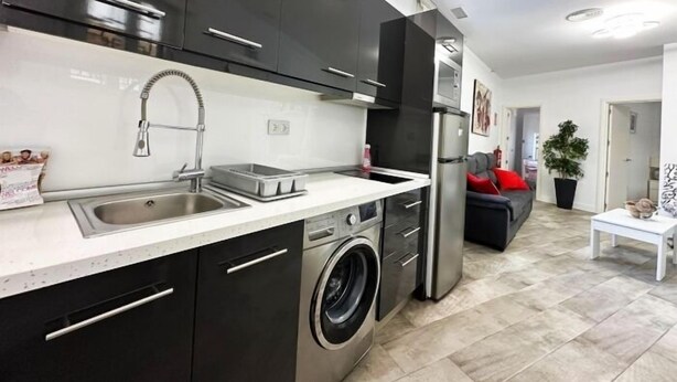 Cozinha moderna com eletrodomésticos de aço inoxidável, máquina de lavar e área de jantar com sofá e mesa.