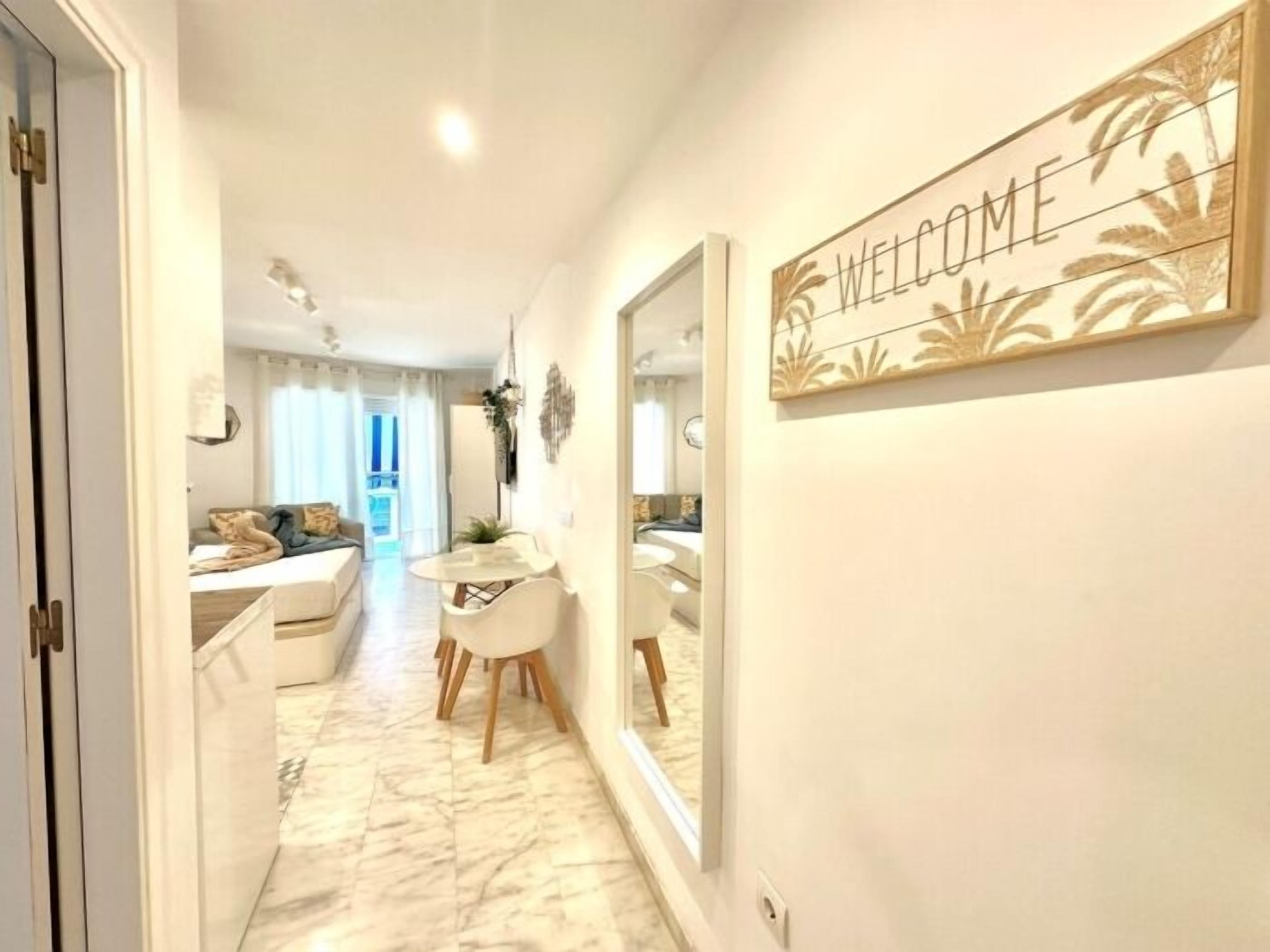 A hallway with a 'WELCOME' sign, a dining area with a table and chairs, and a sofa in the background.