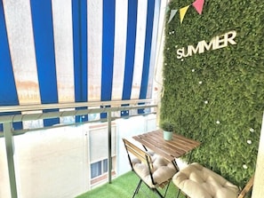 Een klein terras buiten met een tafel en stoelen, een gestreept zonnescherm en een muur met de tekst 'SUMMER'.