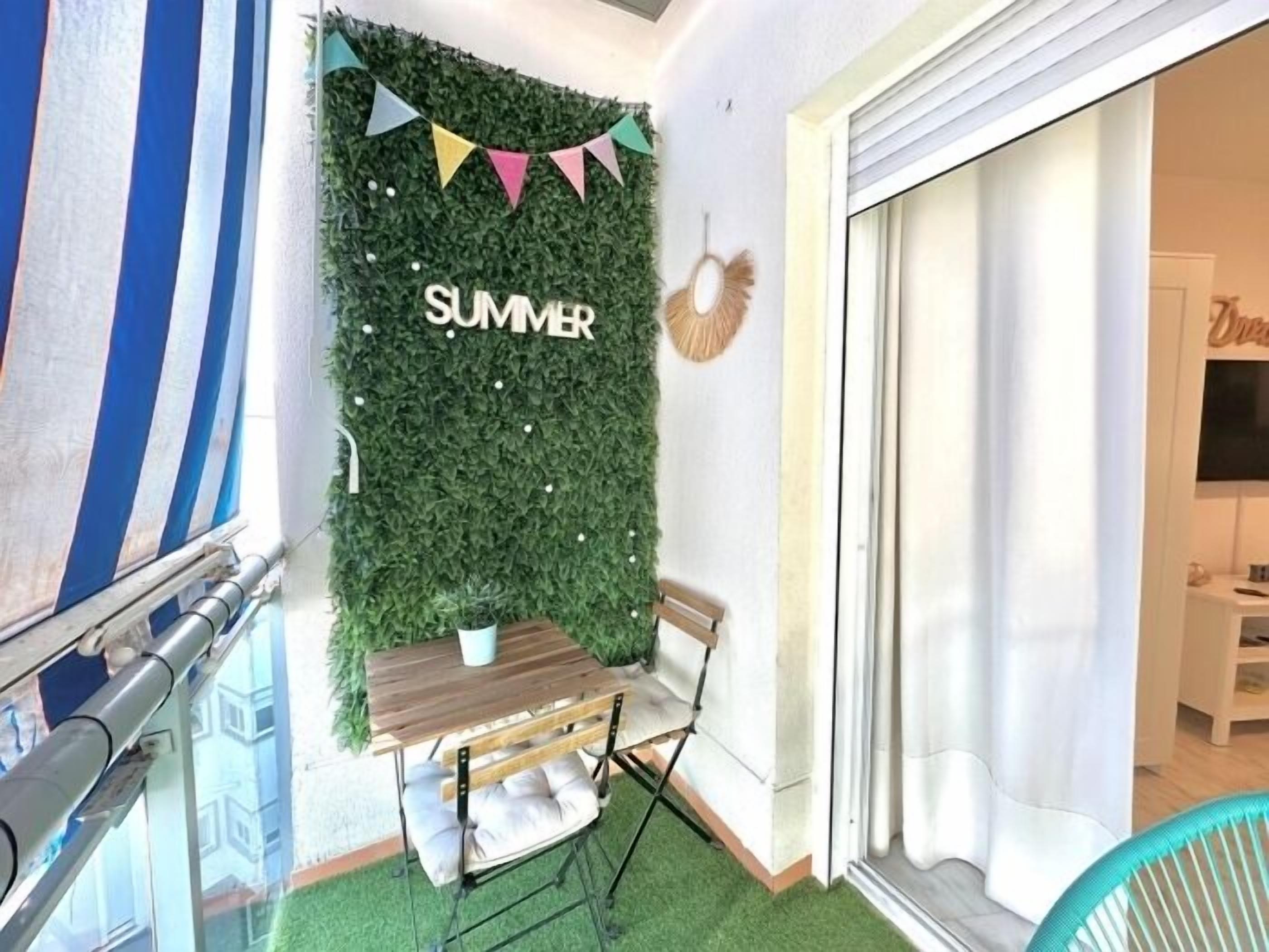 A balcony with a table and chairs, a green artificial turf floor, a wall with a 'SUMMER' sign, and a view of a building through a glass door.