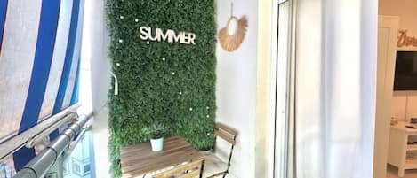 Un balcon avec une table et des chaises, un sol recouvert d’un gazon synthétique vert, un mur orné d’une inscription « SUMMER » et une vue sur un immeuble à travers une porte vitrée.