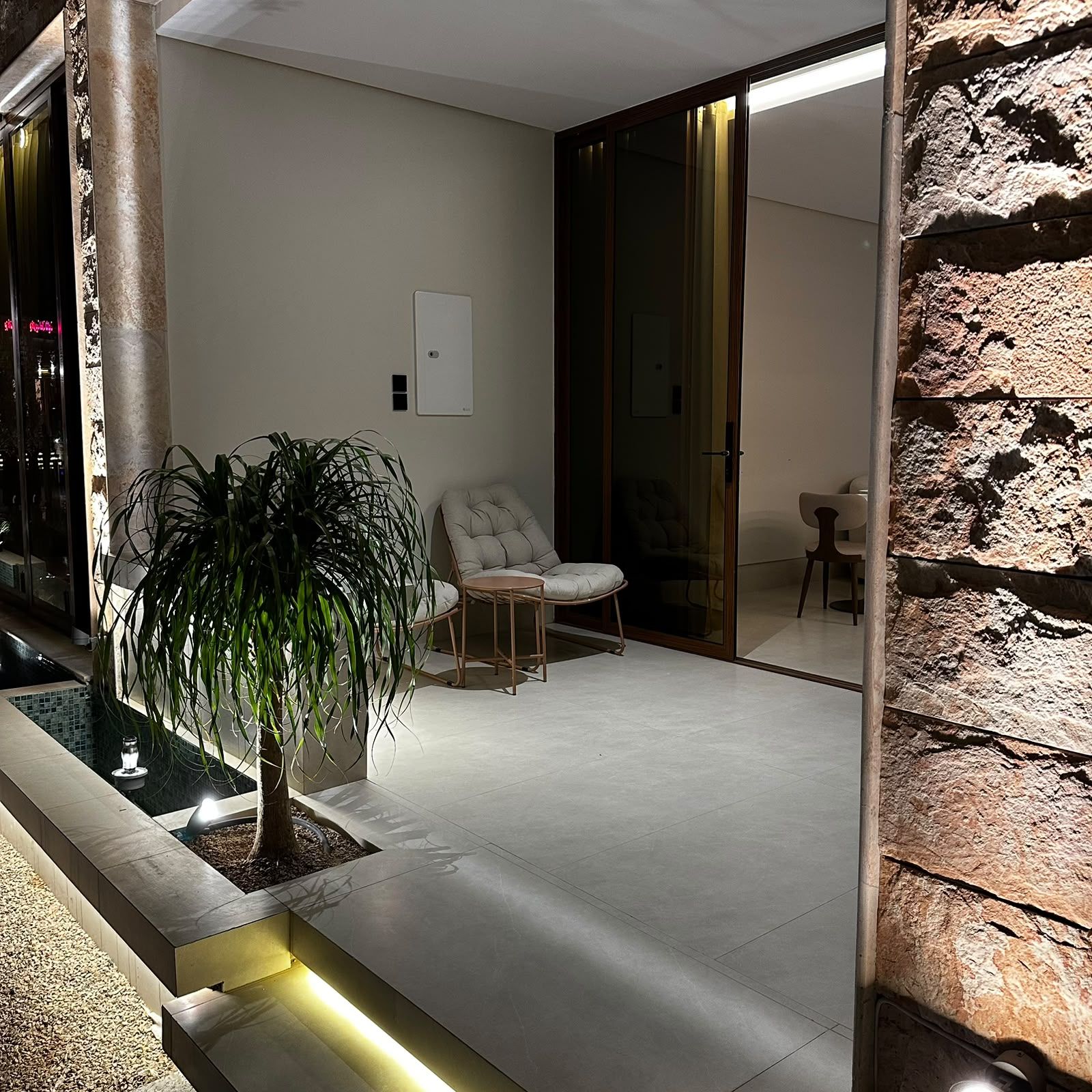A modern outdoor seating area with a potted plant, a cushioned chair, and a glass door leading to a room.