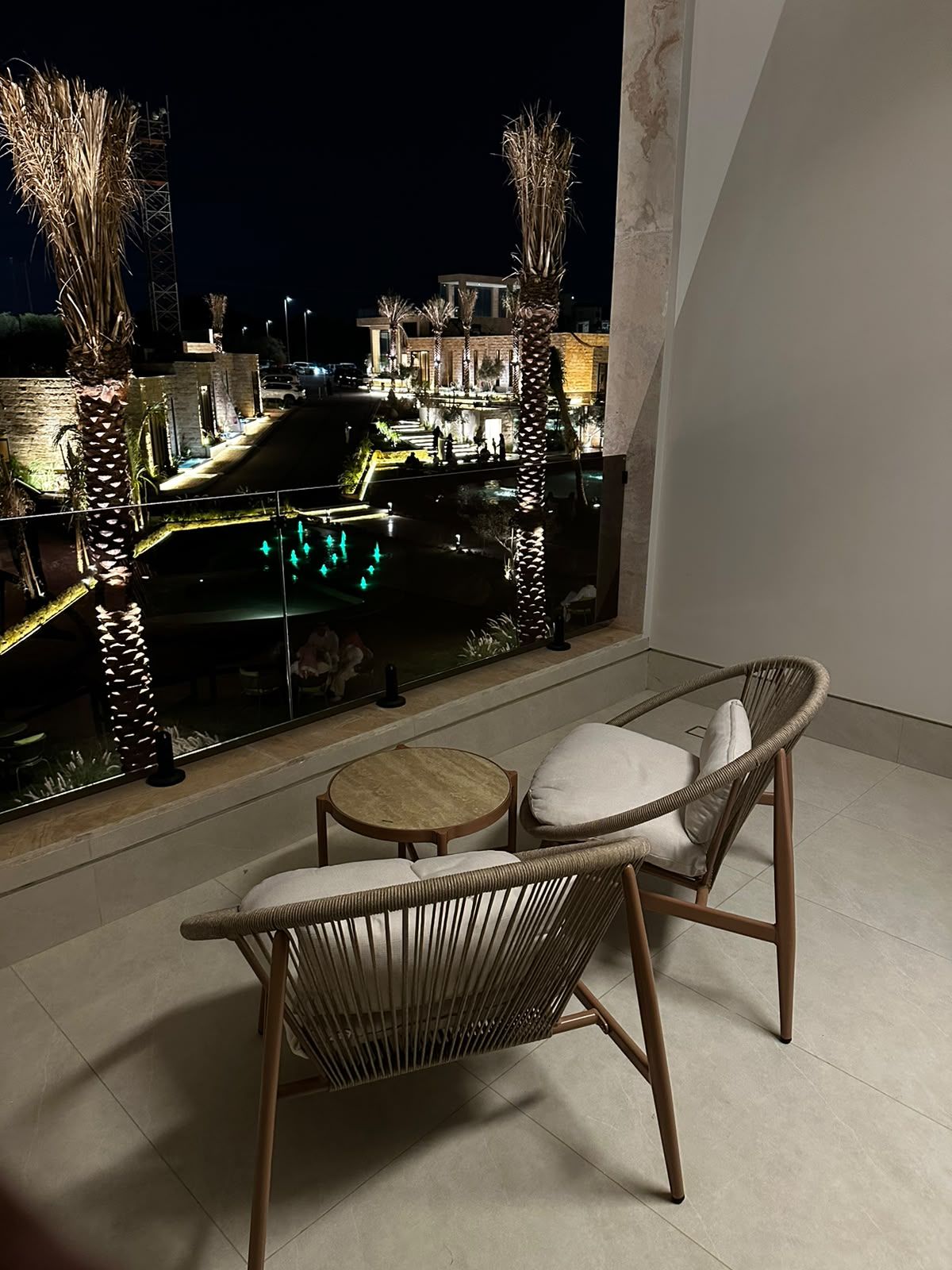 Ein Balkon mit Korbmöbeln, einer Glastrennwand und Blick auf ein beleuchtetes Gebäude bei Nacht.
