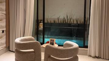A modern room with two round ottomans, a small table with a picture frame, and a view of a swimming pool through a glass partition.