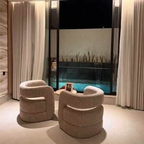 Una habitación moderna con dos otomanos redondos, una mesita con un marco de fotos y vistas a una piscina a través de una pared de vidrio.