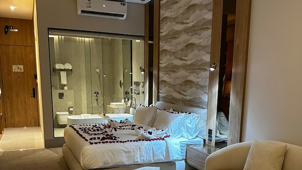 Ein modernes Schlafzimmer mit einem Bett, einem Sofa, einem runden Couchtisch und einem durch eine Glastür einsehbaren Badezimmer.