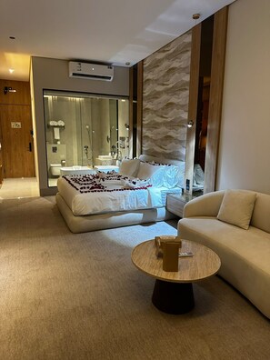 A modern bedroom with a bed, a sofa, a round coffee table, and a bathroom visible through a glass door.