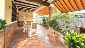 Área de varanda coberta com piso de azulejos, disposição de assentos e plantas em vasos.