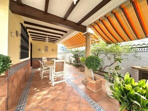 Een overdekt terras met betegelde vloer, een zithoek en potplanten.
