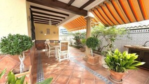 Pátio externo com piso de azulejos, vasos de plantas e área de estar com cadeiras e uma mesa.