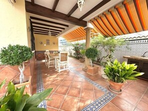 Een terras buiten met betegelde vloer, planten in potten en een zithoek met stoelen en een tafel.