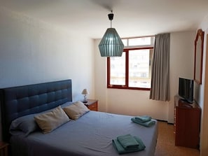 Een slaapkamer met een bed, een nachtkastje, een televisie, een raam met gordijnen en een hanglamp.