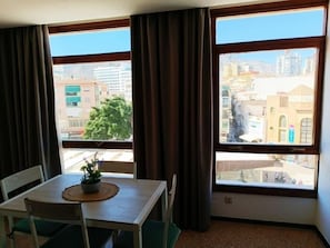 Een kamer met een eettafel en stoelen, grote ramen met gordijnen en uitzicht op gebouwen.