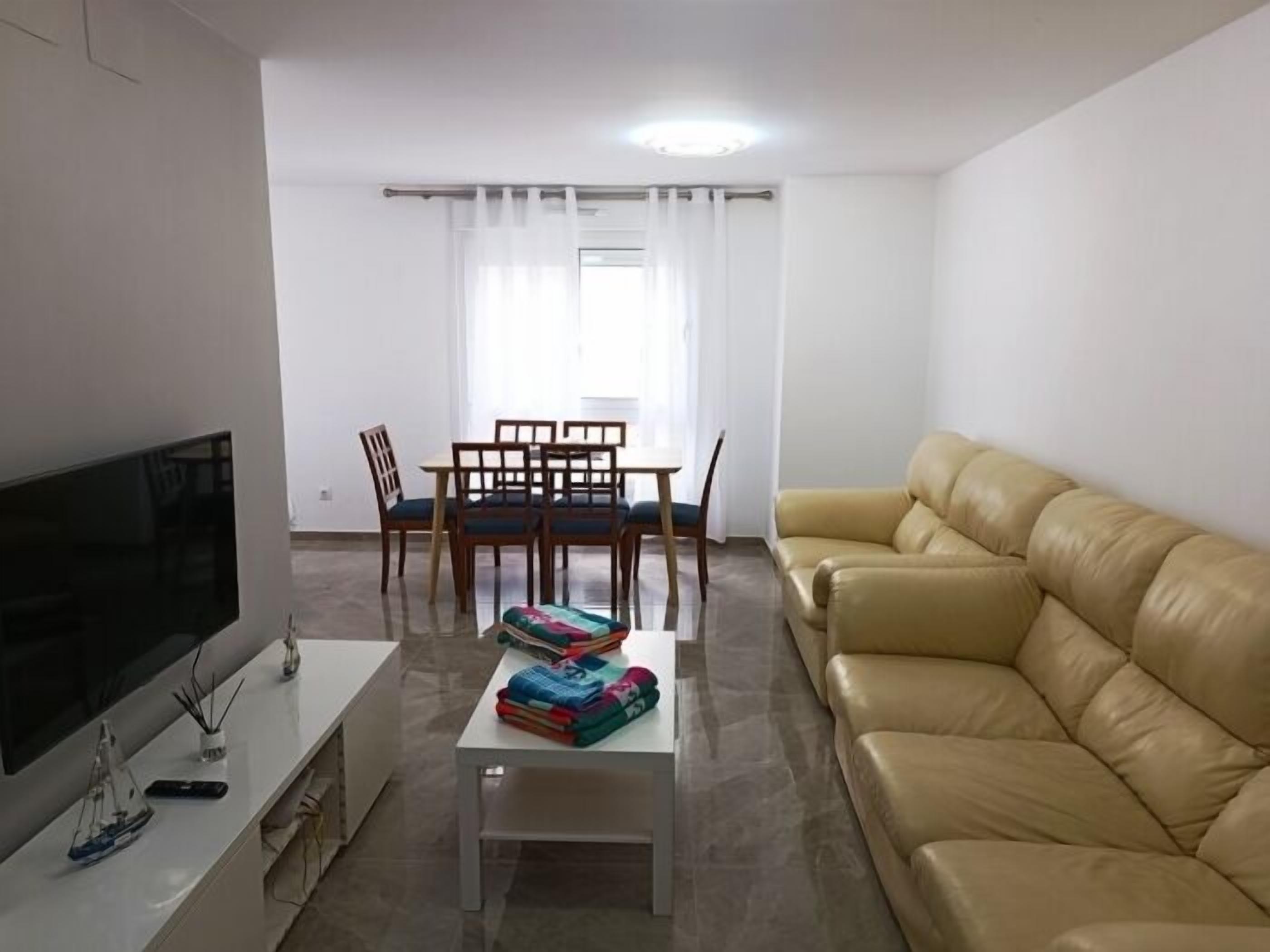 Sala de estar com televisão de tela plana, sofá, mesa de jantar com cadeiras e janela com cortinas.