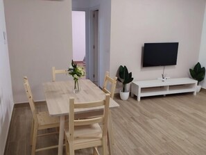Een eethoek met een houten tafel en stoelen, een vaas met bloemen, een flatscreen televisie aan de muur en een witte meubelset.