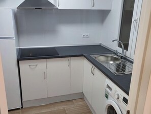 Cozinha compacta com armários brancos, bancada preta, pia de aço inoxidável e máquina de lavar embutida.