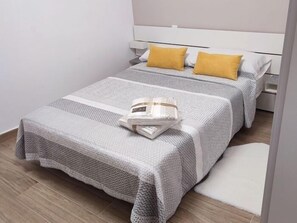 Een tweepersoonsbed met een grijze en witte spreidek, twee gele kussens, en een witte nachtkastje met een lamp.