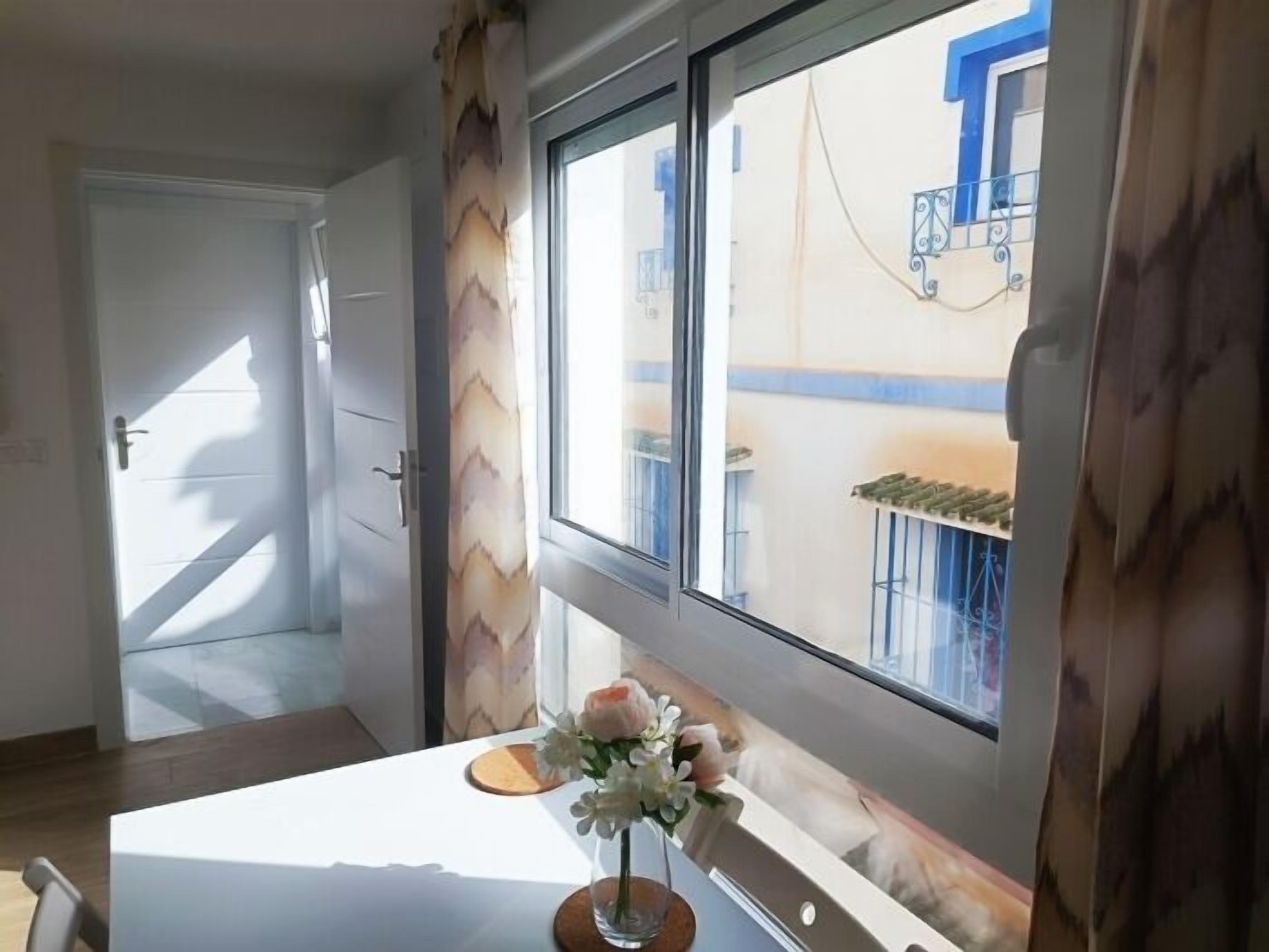 Una habitación con ventana, una mesa blanca con un jarrón de flores y vista a un edificio.