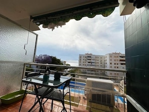 Een balkon met een tafel, stoelen, een groene plantenbak en uitzicht op gebouwen en een zwembad.