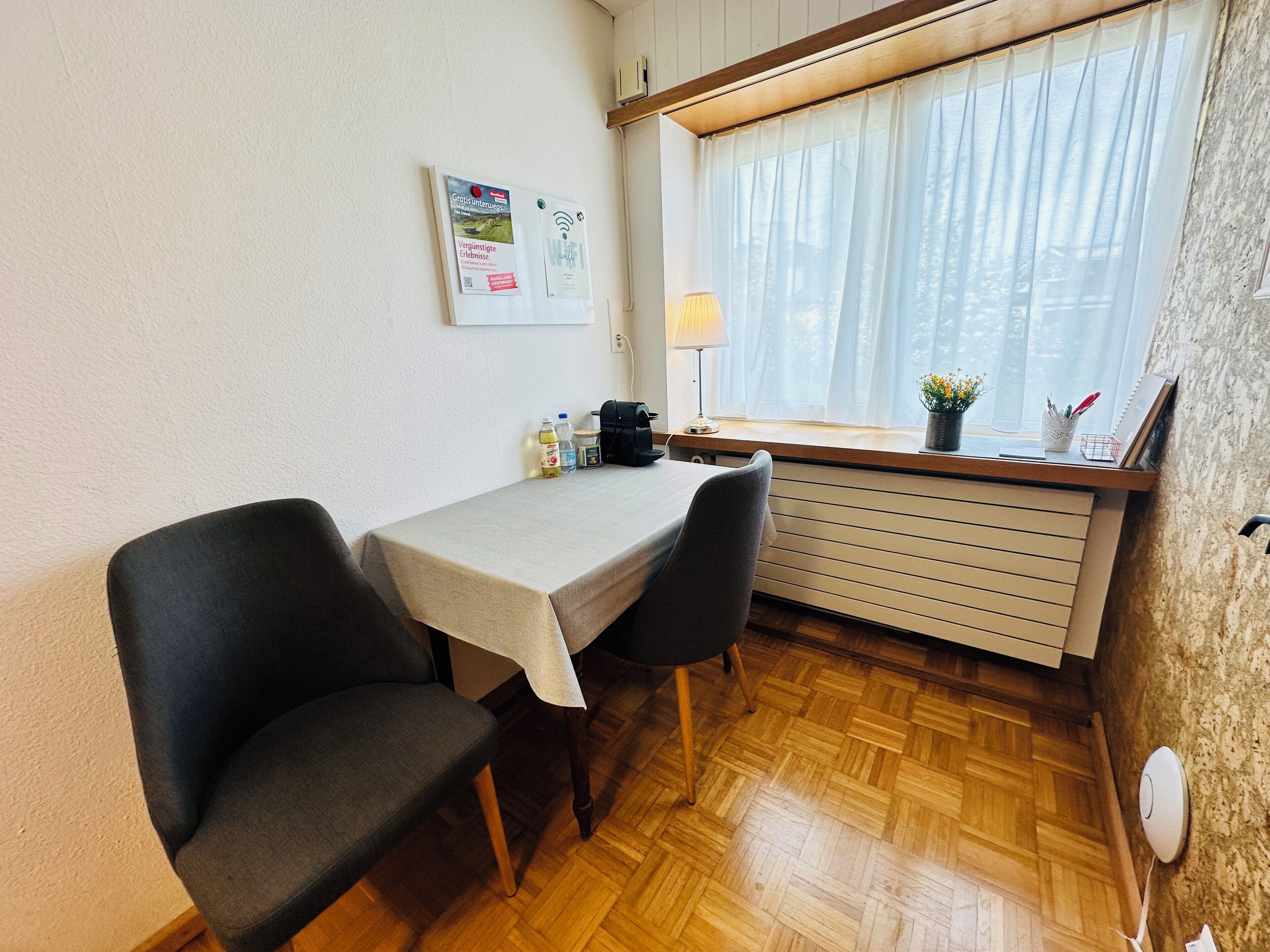 A small, tidy room with a table and two chairs, a window with sheer curtains, a wall-mounted lamp, and a radiator.